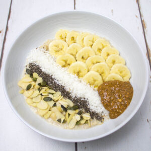 porridge topped with bananas, almonds, grated coconut, almond butter, pumpkin seeds and chia seeds arranged in a white bowl