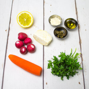 carrot, celeriac, radishes, parsley, lemon, capers and olive oil arranged on a white wooden table
