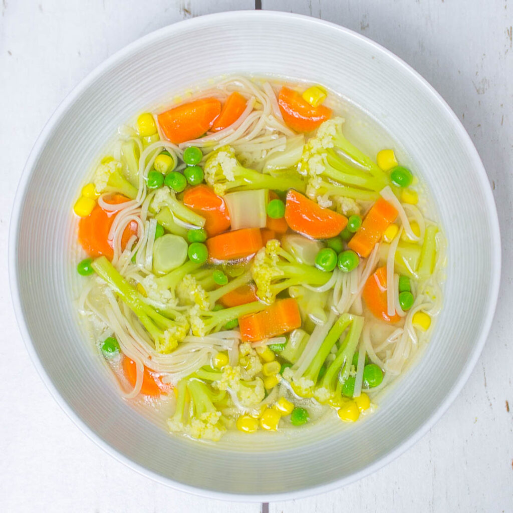 round white bowl filled with carrot, peas, corn and cauliflower soup with brown rice noodles on a white wooden table