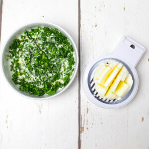 boiled egg in an egg slicer and a mayo with chives in a white bowl, both on a white wooden table