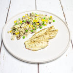 baked white fish and cauliflower rice with red onion, peas and corn arranged on a white round plate