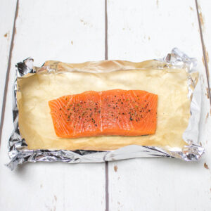 a piece of raw salmon seasoned with salt and pepper on a baking paper inside a tin foil tray
