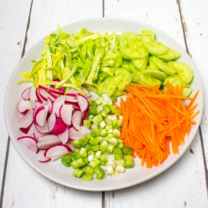 shredded cabbage, chopped cucumber, radishes and salad onions and thinly sliced carrots arranged on a white plate