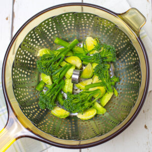 chopped tenderstem broccoli and brussels sprouts in a steel steaming basket in a glass saucepan
