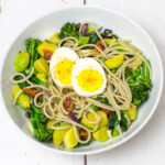 white bowl with cooked brown rice spaghetti mixed with lots of steamed vegetables and sundried tomatoes topped with a boiled egg chopped in half